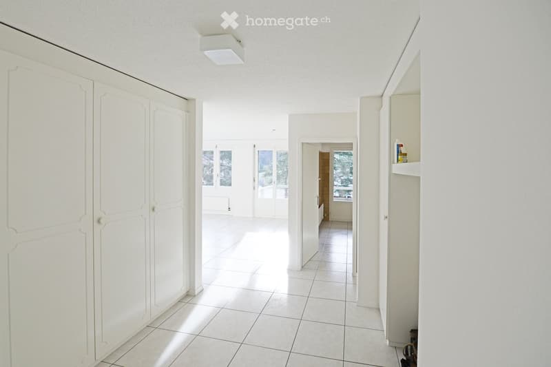 Bright, clean hallway with white tiled flooring, white walls, and built-in storage units on both sides. Natural light streams in from the end of the hallway through large windows.
