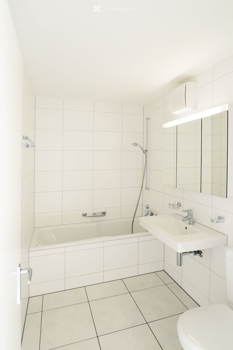 Modern bathroom with white tiled walls and floor, featuring a bathtub, sink, mirror, and toilet.