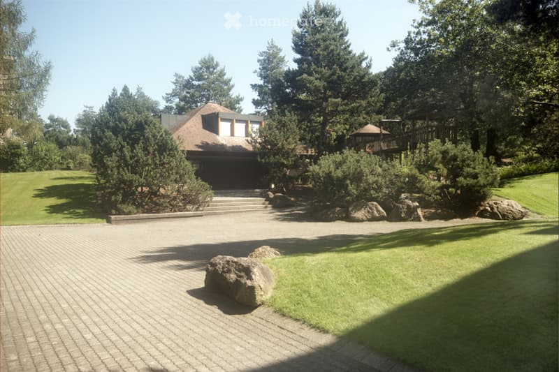 Well-maintained garden with a large house in the background, featuring a paved driveway, manicured lawn, trees, and decorative rocks.