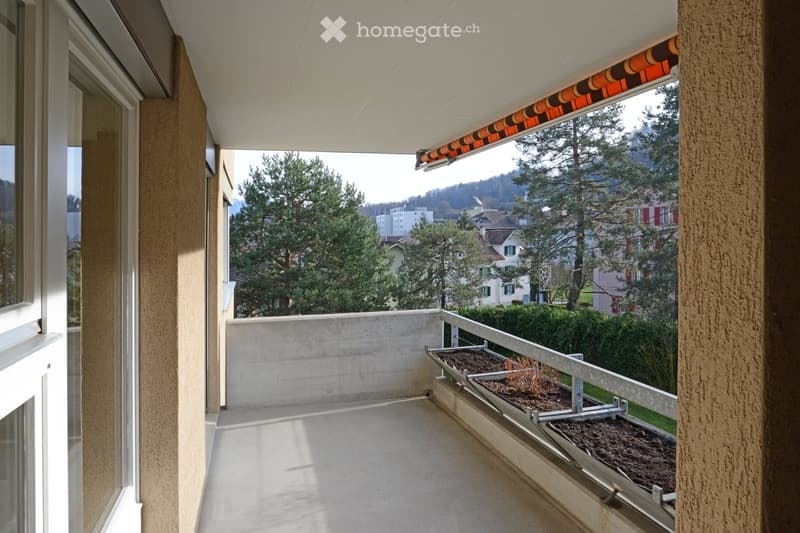 Balcony with a concrete floor, white ceiling, and a retractable awning. The balcony overlooks a green area with trees and houses in the background.