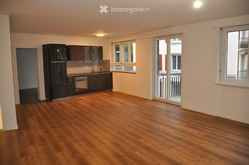 Empty room with wooden flooring, white walls, and a modern kitchen area with cabinets and a sink. Features large windows and a sliding glass door leading to a balcony.
