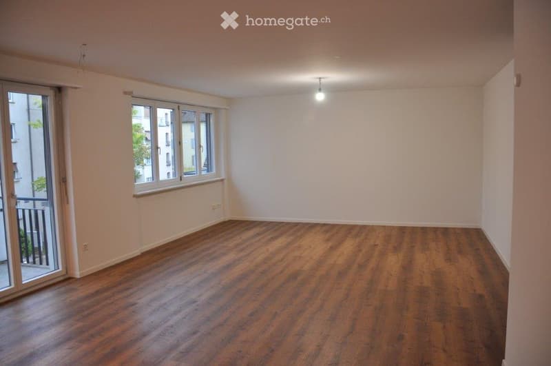 Empty room with wooden flooring, white walls, large windows, and a sliding glass door leading to a balcony.