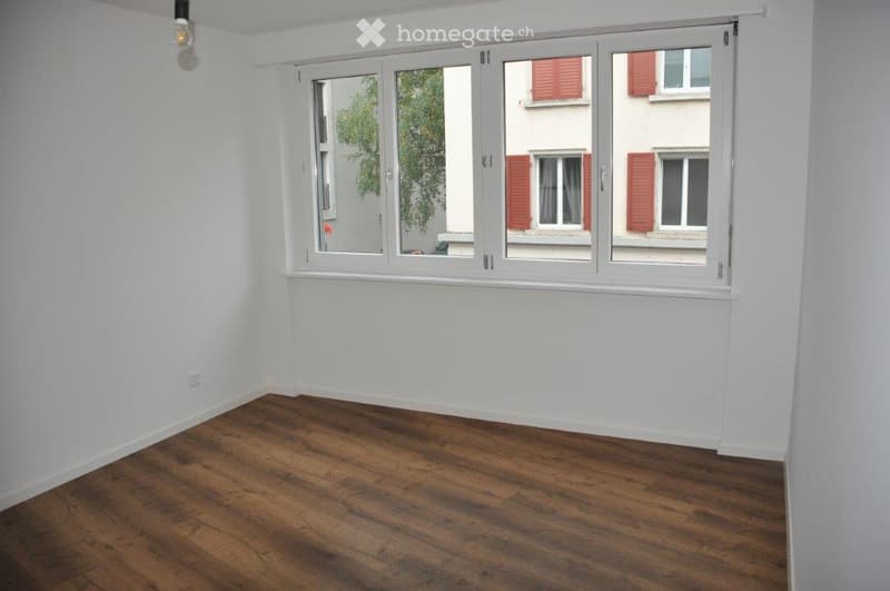 Empty room with white walls, wooden floor, and large windows showing a view of neighboring buildings with red shutters.