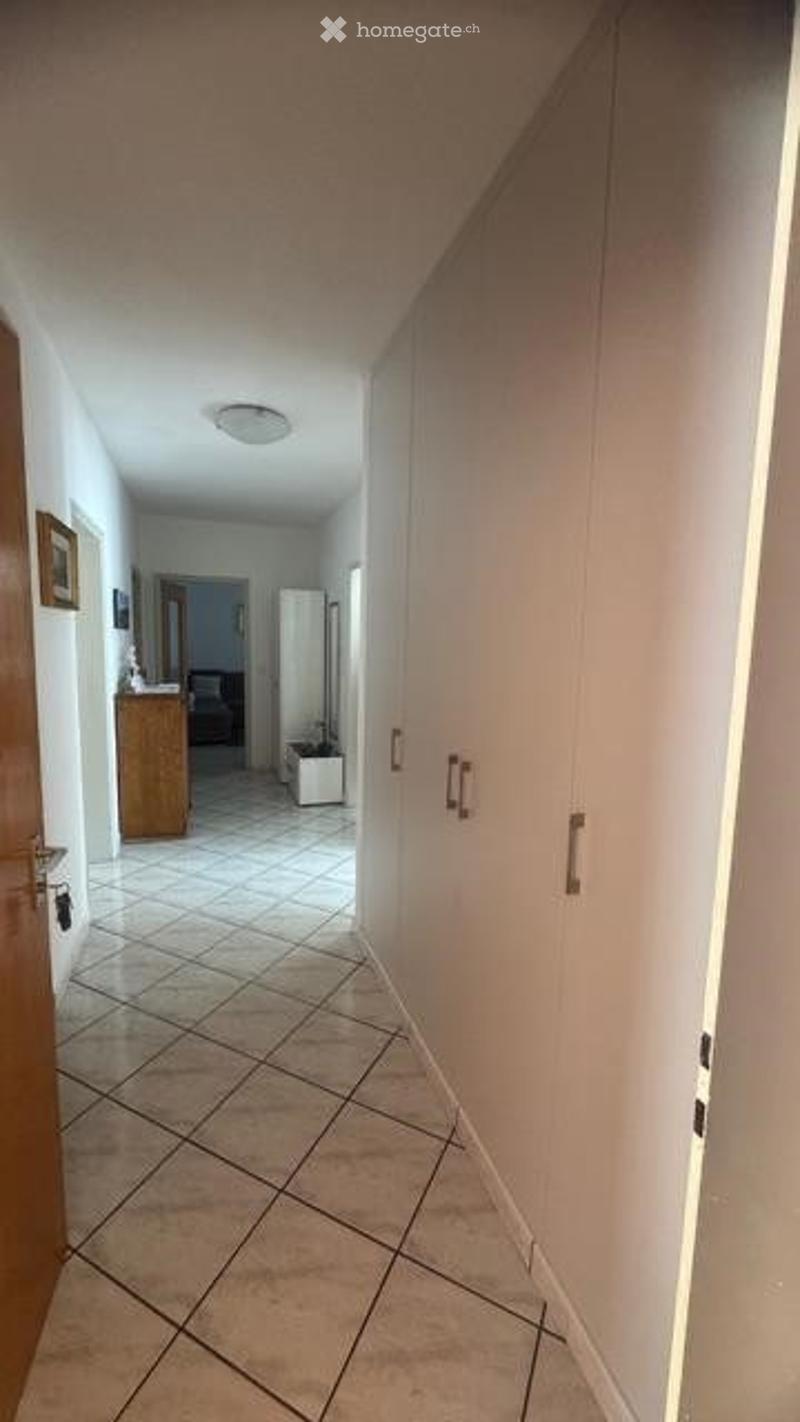 A clean, tiled hallway with white walls, a wooden cabinet, and a small room visible at the end.