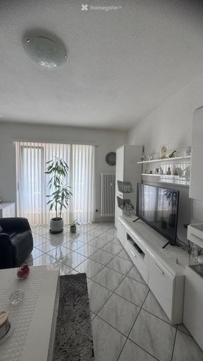 Modern living room with white walls, tiled floor, large window with blinds, black leather sofa, coffee table, flat-screen TV on white stand, plant, and shelf with decorative items.