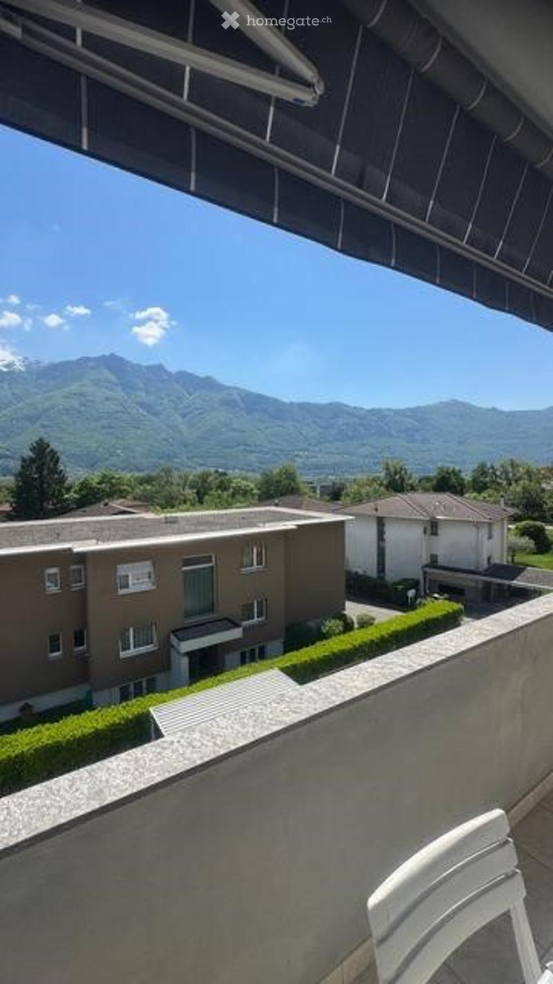 Balcony with a view of a residential area and mountains in the background. A white plastic chair is visible on the balcony.