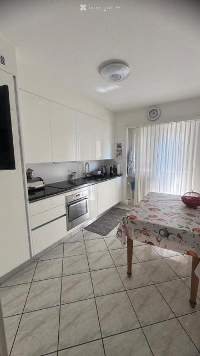 Modern kitchen with white cabinets, stainless steel appliances, and a tiled floor. A small table with a floral tablecloth and a bowl is visible.