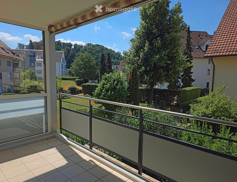 Balcony with tiled floor, glass railing, overlooking a green landscape with trees and houses.