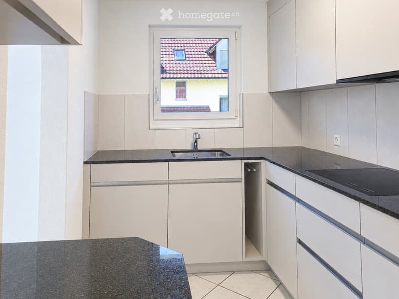 Modern kitchen with white cabinets, dark granite countertops, a stainless steel sink, and an induction cooktop. A window offers a view of a tiled roof.