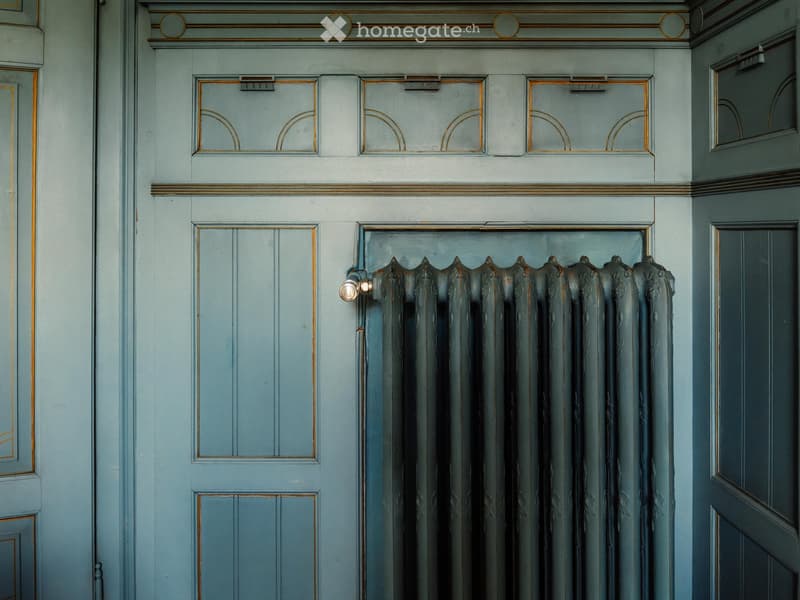 A hallway with light blue walls, ornate gold-framed doors, and a decorative radiator with a vintage design.