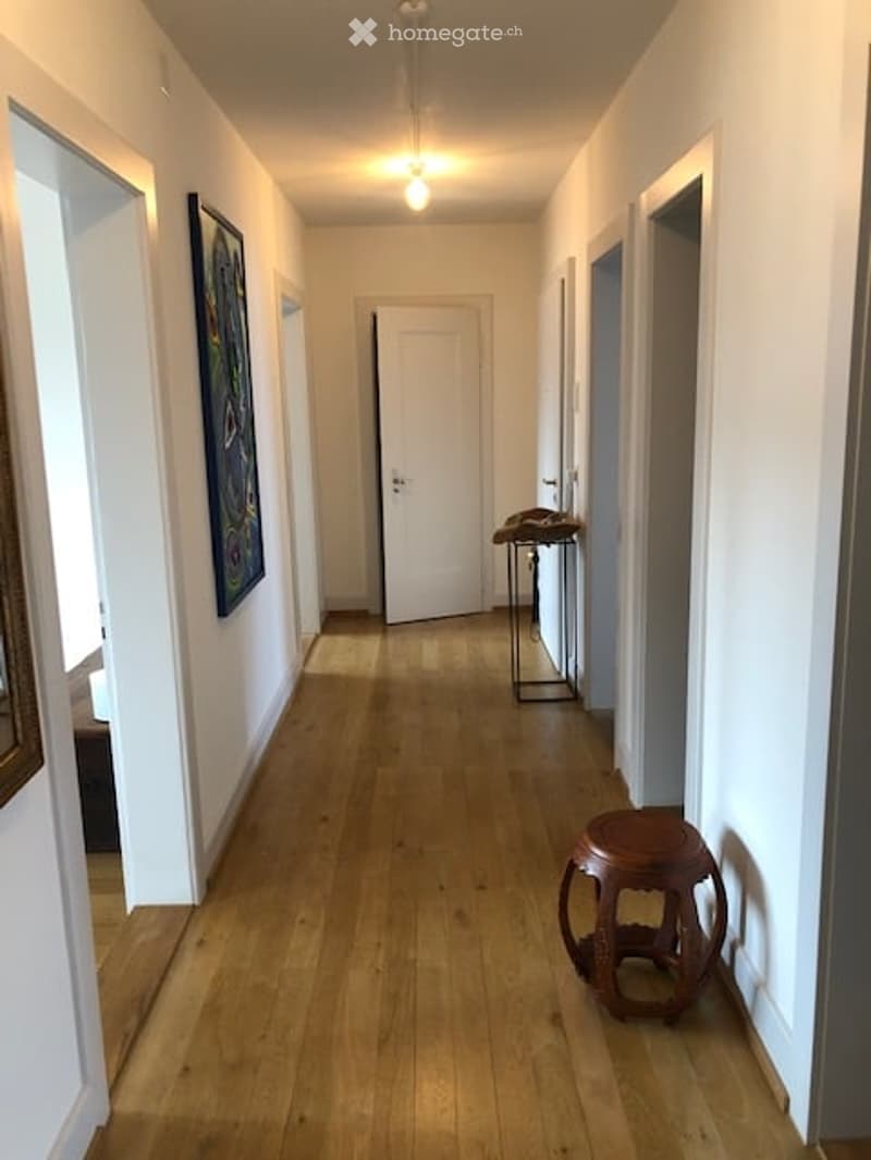 A long, narrow hallway with wooden flooring, white walls, and a single light fixture on the ceiling. There are two open doorways at each end, and a small stool and a small table are placed against the right wall.