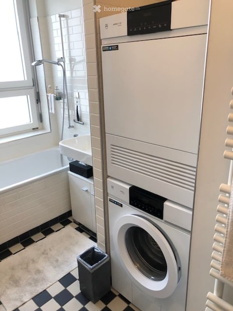 Modern bathroom with a bathtub, sink, shower, and stacked washing machine/dryer unit. The floor has a black and white checkered pattern, and there is a small trash bin near the washing machine.