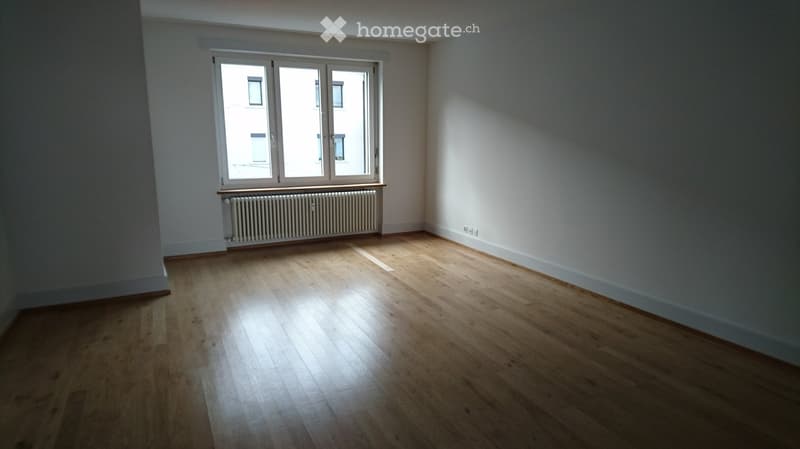 Empty room with wooden flooring, white walls, and large windows allowing natural light to enter. A radiator is visible beneath the windows.