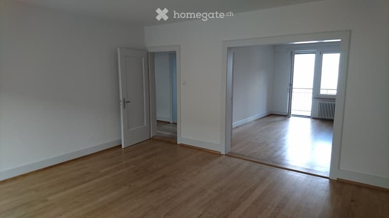 Empty room with wooden flooring, white walls, and open doors leading to another room and a balcony area.