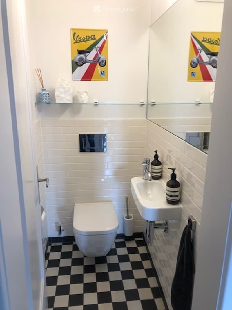 Modern bathroom with white tiles, black and white checkered floor, a toilet, a sink with a mirror, a towel, and decorative posters on the wall.