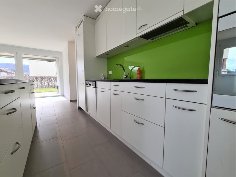 Modern kitchen with white cabinets, tiled floor, and a green accent wall. Features a sink with a faucet, a dishwasher, and a bottle on the counter.