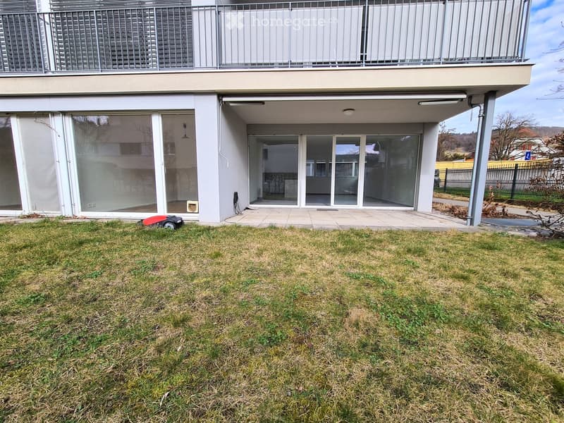 Lawn area in front of a modern house with large glass sliding doors, a balcony above, and a small patio area.