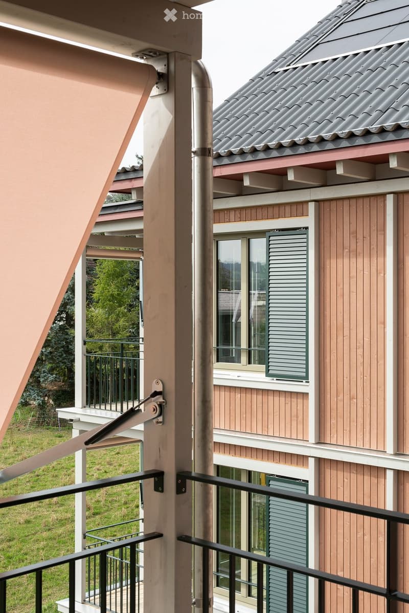 Balcony with black metal railing, beige awning, and view of a wooden-sided building with windows and shutters.