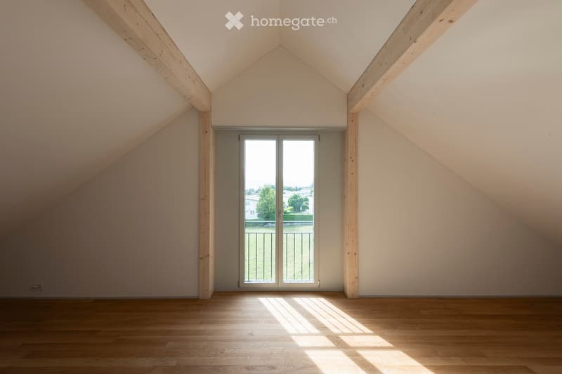 Empty attic room with wooden floors, slanted ceilings, and a large window offering a view of the outdoors.
