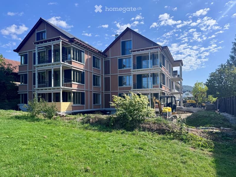 Modern two-story house with red-brown facade, large windows, and balconies, surrounded by green lawn and construction equipment.