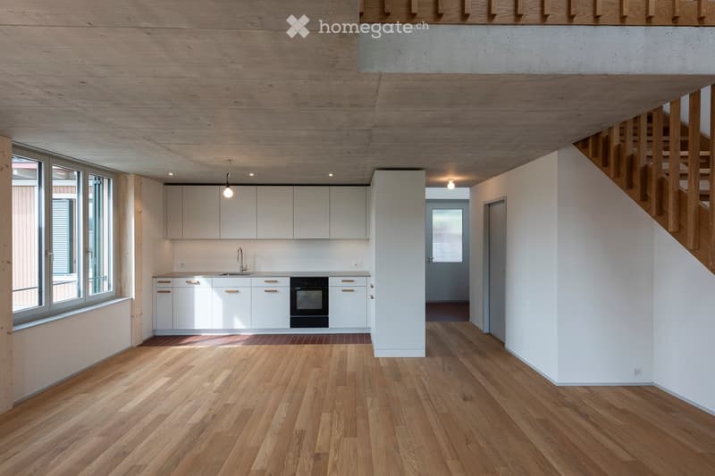 Open-plan kitchen and living area with light wood flooring, white cabinets, and a modern oven. The space features large windows and a wooden staircase leading to an upper level.