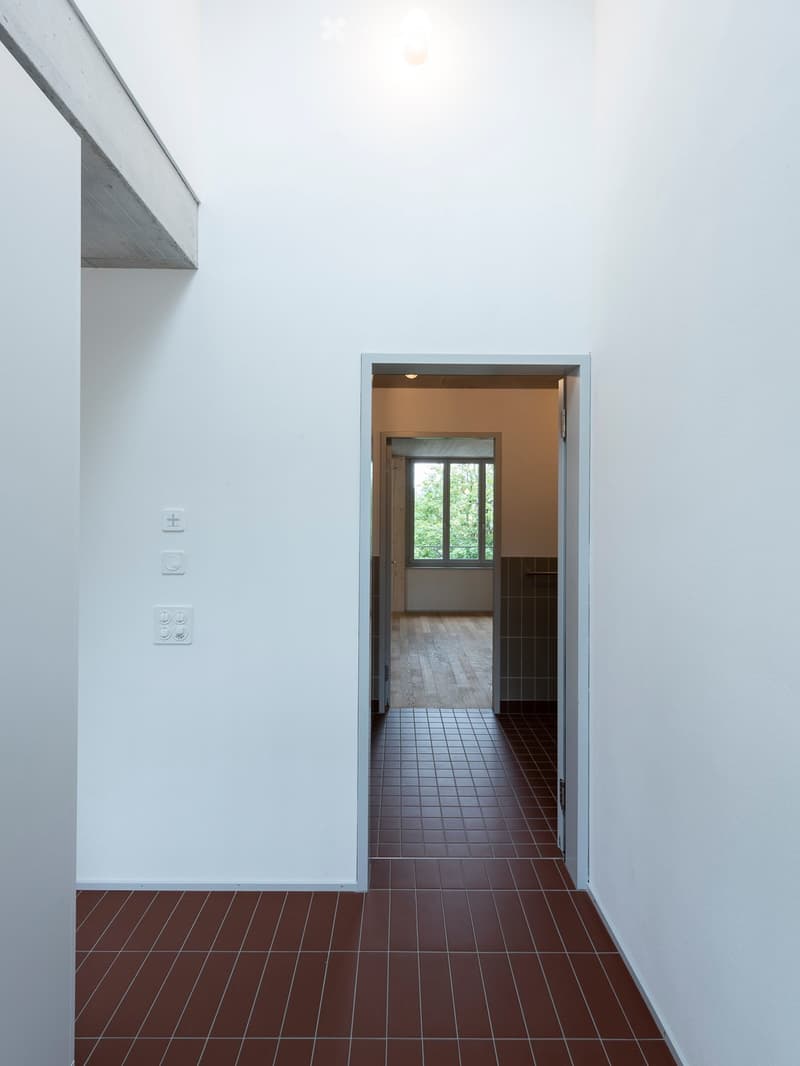 Minimalist hallway with red tiled floor, white walls, and recessed lighting. Features a doorway leading to another room with wooden flooring and large windows.