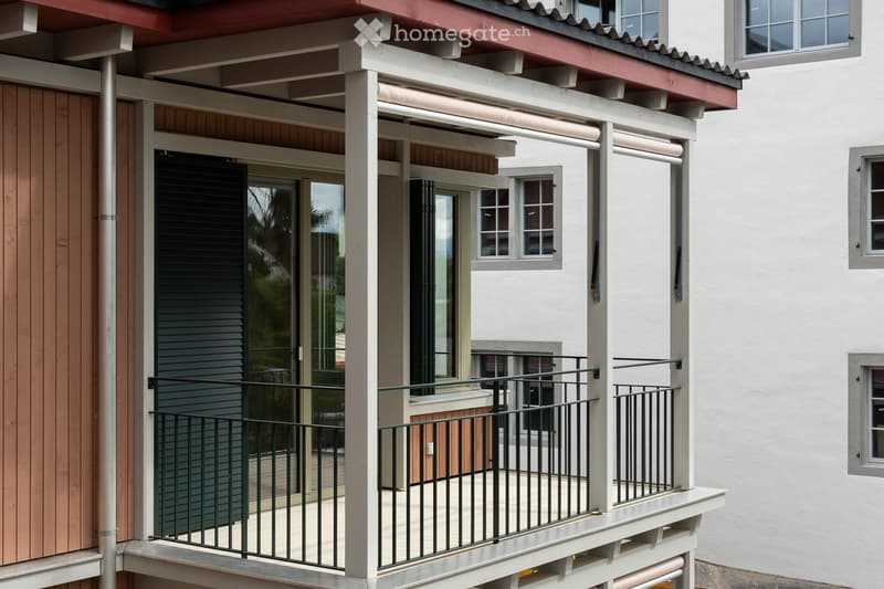 Balcony with metal railing, glass doors, and wooden decking, attached to a multi-story building with white walls and multiple windows.