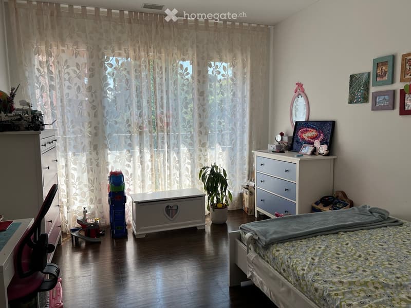 A well-lit bedroom with a bed, a chair, a plant, a mirror, picture frames, and toys. The room features wooden flooring and large windows with curtains.