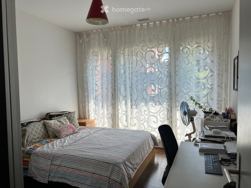 A well-lit bedroom with a bed, nightstand, desk, chair, fan, and laptop. The room features large windows with patterned curtains and a modern lamp hanging from the ceiling.