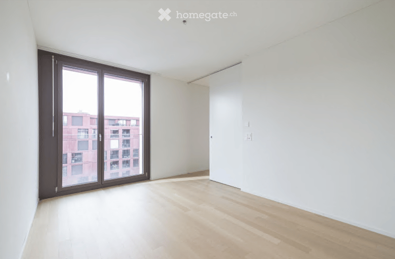 Empty room with light wooden flooring, white walls, and a large glass sliding door leading to a view of a building outside.
