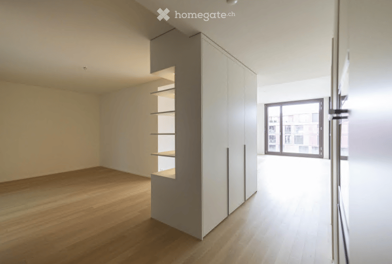 Empty room with light wooden flooring, white walls, built-in white cabinetry with open shelving, and a large window showing a cityscape view.