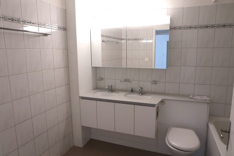 Modern bathroom with white tiles, double sink, mirrored cabinet, toilet, and bathtub.