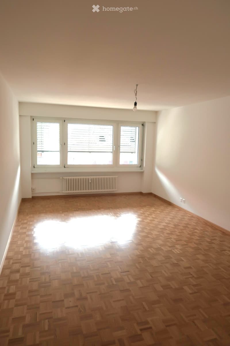 Empty room with wooden parquet flooring, white walls, three windows with blinds, radiator, and a single ceiling light fixture.