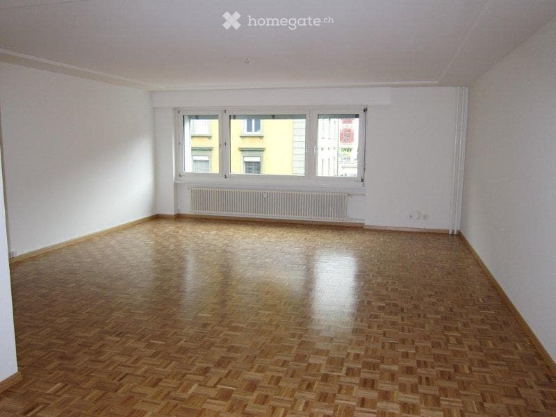 Empty room with wooden flooring, white walls, and large windows with a radiator below. No furniture or equipment present.