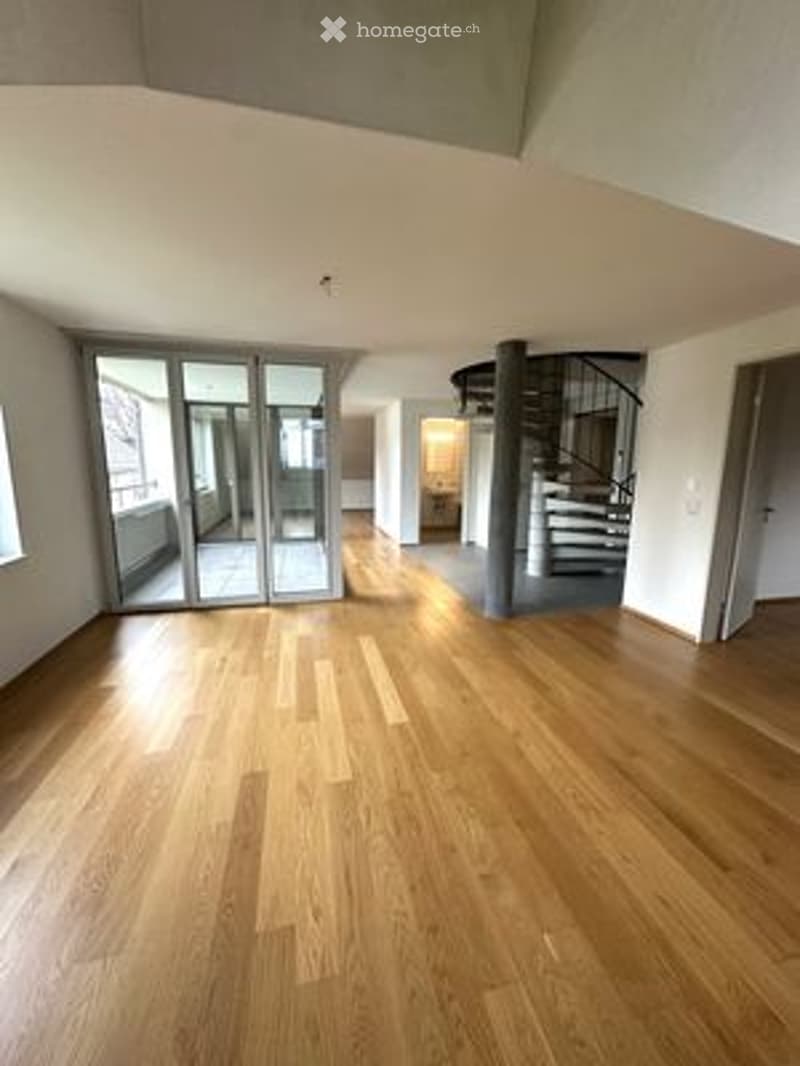 Bright, empty room with wooden flooring, large glass sliding doors, and a visible staircase in the background.