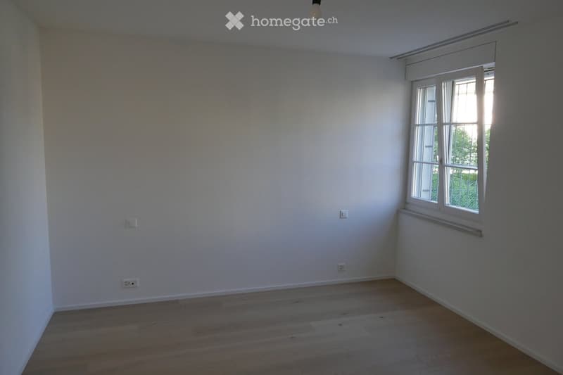 Empty room with white walls, wooden flooring, and a large window with white frames. No furniture or equipment is present.