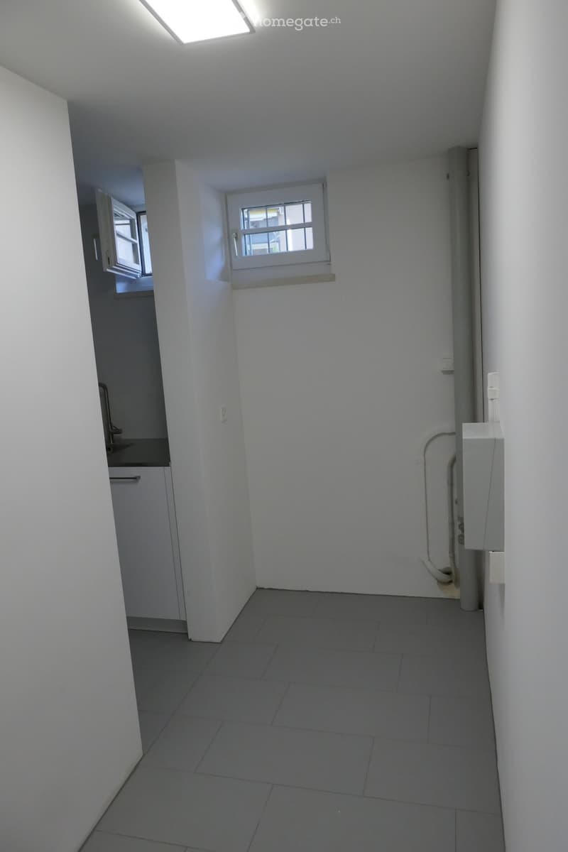 Empty room with white walls, grey tiled floor, small window, and a small sink area visible in the background.