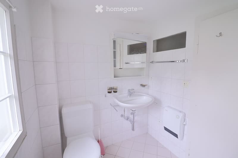 A small, clean bathroom with white tiles, a toilet, a sink with a mirror above it, and a towel rack on the wall.