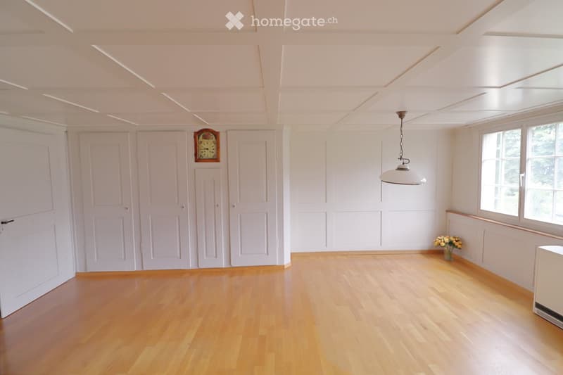 Empty room with wooden flooring, white walls, and large windows. Features a hanging light fixture and a small vase with flowers on the windowsill.