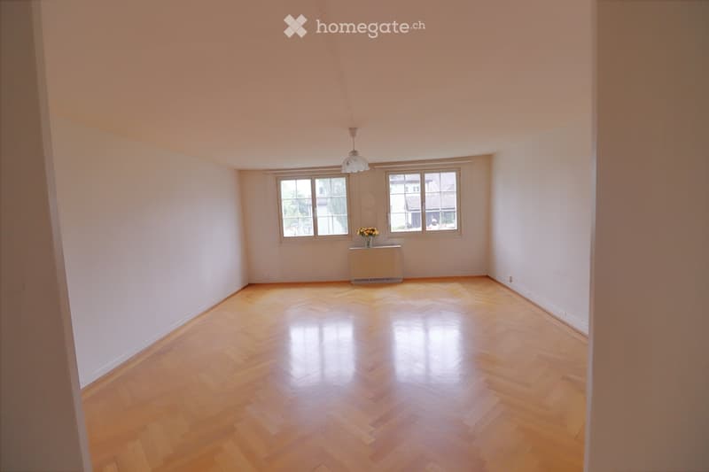 Empty room with wooden herringbone flooring, white walls, two windows, and a radiator with a small flower arrangement on top.