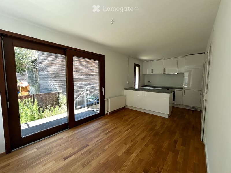 Open-plan space with wooden flooring, large sliding glass door leading to outdoor area, modern kitchen with white cabinets, sink, refrigerator, and oven.