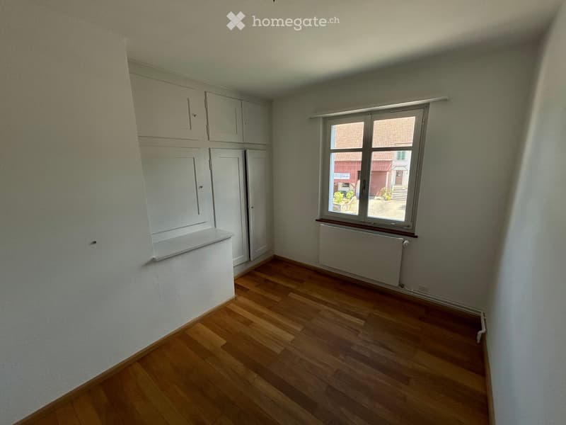 Empty room with wooden flooring, white walls, built-in cabinets, a window with blinds, and a radiator below the window.