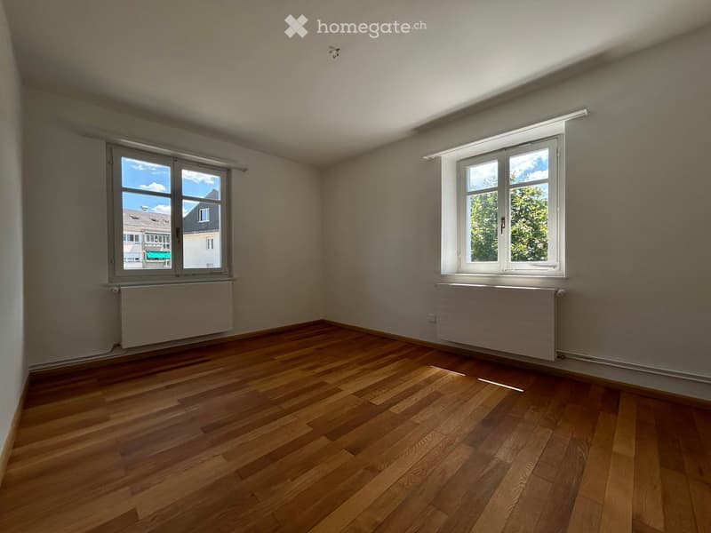 Empty room with wooden flooring, two windows, and radiators beneath them.