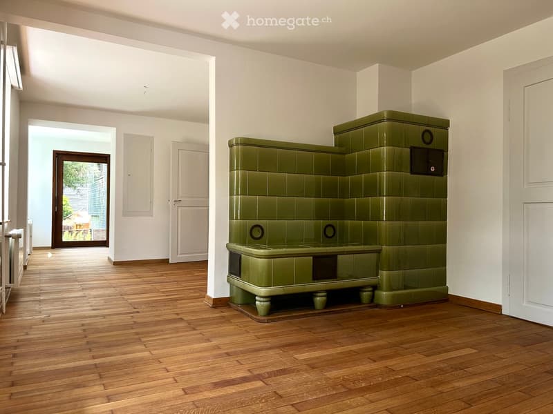 A spacious room with wooden flooring and white walls, featuring a large green tiled fireplace as the central focal point. The room has large windows and doors leading to an outdoor area.