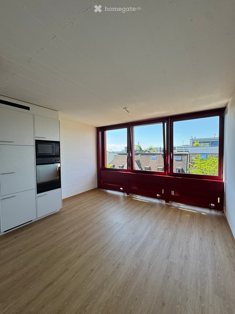 Empty room with wooden flooring, white cabinets, and large windows offering a city view.