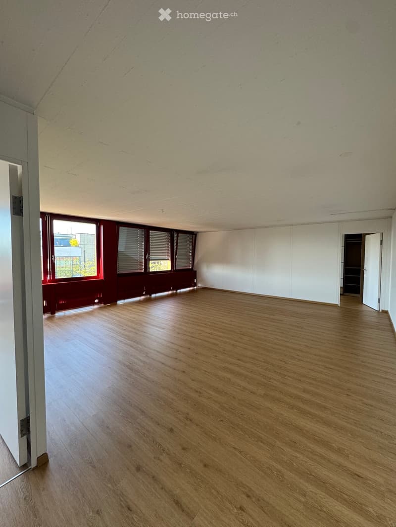 Empty room with wooden flooring, white walls, large windows with blinds, and a door leading to another space.