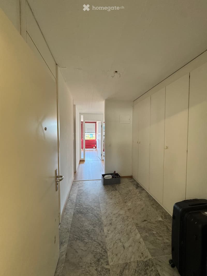 A narrow hallway with marble flooring, white walls, and a small black suitcase placed on the right side. The hallway leads to an open doorway with a red door visible in the background.