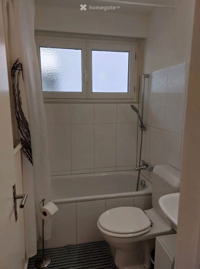 Small bathroom with a toilet, sink, bathtub, and shower. Features tiled walls, a window, and a toilet paper holder.