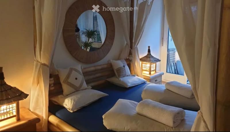 A cozy bedroom with a large bed, multiple pillows, neatly rolled towels, a nightstand with a lamp, and a round wooden-framed mirror on the wall.
