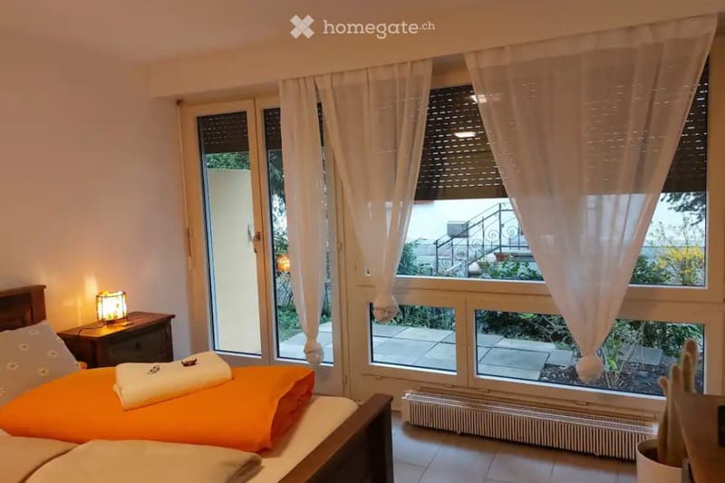 Bright bedroom with large windows and a balcony view, featuring a double bed with orange bedding, a nightstand with a lamp, and sheer white curtains.